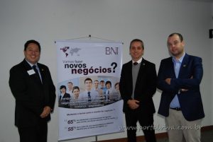 três homens de terno ao lado de faixa com os dizeres "Vamos fazer novos negócios?" durante jantar do BNI