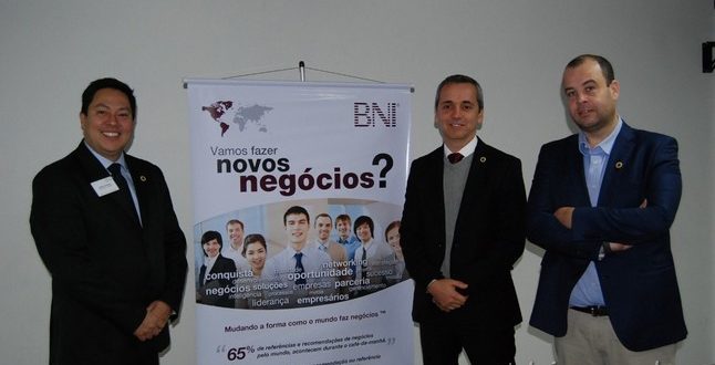 três homens de terno ao lado de faixa com os dizeres "Vamos fazer novos negócios?" durante jantar do BNI