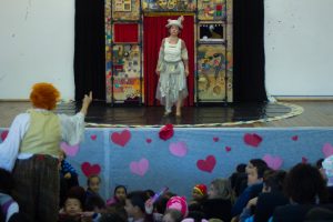 Uma atriz se apresenta no palco do CEUC, em Caucaia do Alto, Cotia, com um pequeno cenário ao fundo, enquanto seu colega se apresenta em meio às crianças que compõem o público