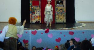 Uma atriz se apresenta no palco do CEUC, em Caucaia do Alto, Cotia, com um pequeno cenário ao fundo, enquanto seu colega se apresenta em meio às crianças que compõem o público