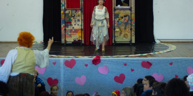 Uma atriz se apresenta no palco do CEUC, em Caucaia do Alto, Cotia, com um pequeno cenário ao fundo, enquanto seu colega se apresenta em meio às crianças que compõem o público