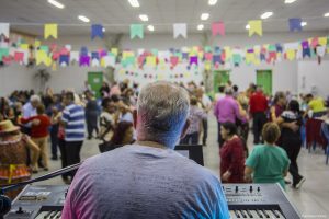 um tecladista visto de costas com o público à sua frente, em segundo plano, durante baile junino no Espaço Movimenta Cotia