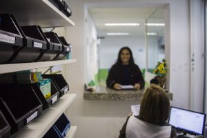 uma mulher sorri e olha para a câmera enquanto outra a atende na farmácia de alguma UBS de Cotia