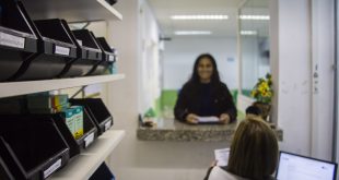 uma mulher sorri e olha para a câmera enquanto outra a atende na farmácia de alguma UBS de Cotia