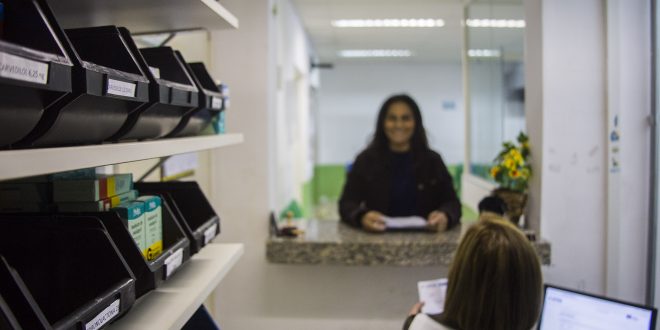 uma mulher sorri e olha para a câmera enquanto outra a atende na farmácia de alguma UBS de Cotia