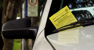 detalhe do parabrisa de um beículo com uma notificação de zona azul presa no limpador e uma placa de sinalização de zona azuil ao fundo, em desfoque