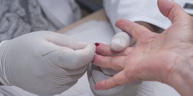 close da mão de uma pessoa tendo uma gota de sangue extraída por duas mãos médicas