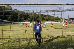 um goleiro visto de costas e através da rede do fundo do gol enquanto uma partida de futebol acontece mais ao fundo