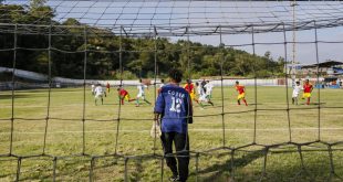 um goleiro visto de costas e através da rede do fundo do gol enquanto uma partida de futebol acontece mais ao fundo