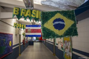 um corredor de uma escola pública de Cotia com bandeiras das nações participantes penduradas e seus respectivos nomes ao lado. A bandeira do Brasil aparece em primeiro plano, à direita, coom a bandeira da Costa Rica atrás, à esquerda, e parte da bandeira de Portugal