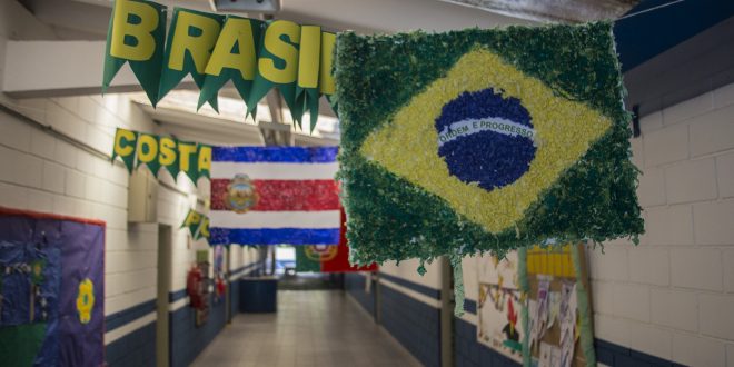 um corredor de uma escola pública de Cotia com bandeiras das nações participantes penduradas e seus respectivos nomes ao lado. A bandeira do Brasil aparece em primeiro plano, à direita, coom a bandeira da Costa Rica atrás, à esquerda, e parte da bandeira de Portugal