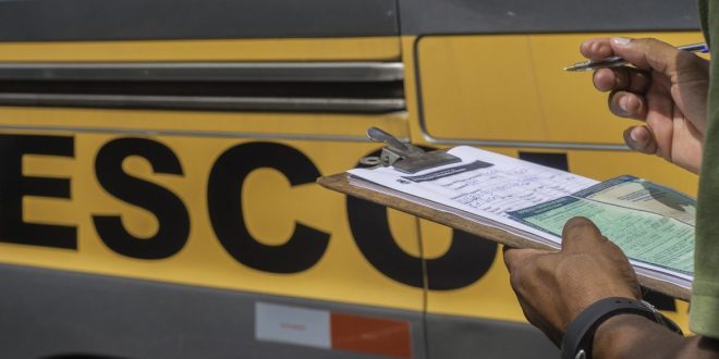 close das mãos de um funcionário da Settrans vistoriando uma vã escolar com uma prancheta