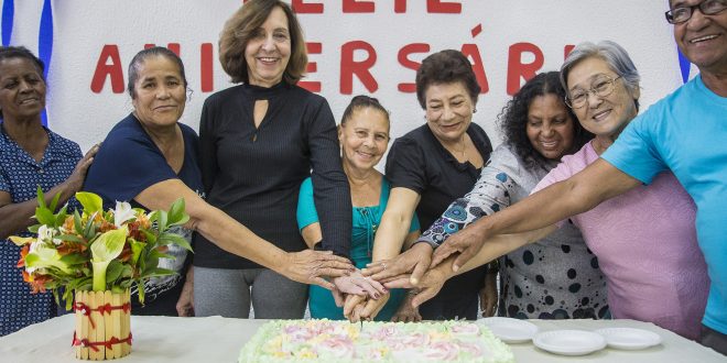 vários aniversariantes do mês partem um mesmo pedaço de bolo enquanto sorriem para a câmera