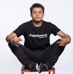 emerson ceará sentado em uma cadeira e fazendo uma careta ante um fundo totalmente branco e com uma camiseta escrito '#aquitransa - @emersonceara'