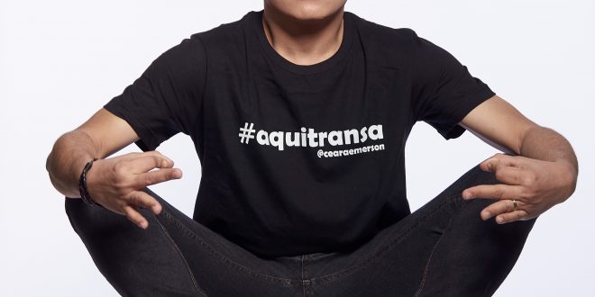 emerson ceará sentado em uma cadeira e fazendo uma careta ante um fundo totalmente branco e com uma camiseta escrito '#aquitransa - @emersonceara'