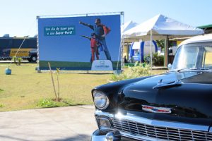 foto parcial da parte da frente de um carro dos anos 50/60 com um outdoor ao fundo com a imagem de um garoto e seu pai logo atrás com uma capa de superherói e os dizeres 'um dia de lazer para o seu super-herói'