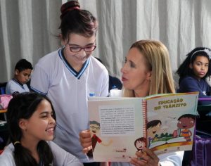 uma professora conversa com duas alunas segurando um livro com o título 'Educação no Trânsito'