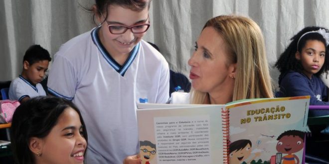 uma professora conversa com duas alunas segurando um livro com o título 'Educação no Trânsito'