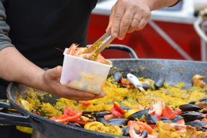 uma pequena porção de paella sendo colocada em um pote, com a grande panela de paella vista logo atrás