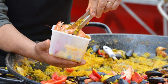 uma pequena porção de paella sendo colocada em um pote, com a grande panela de paella vista logo atrás