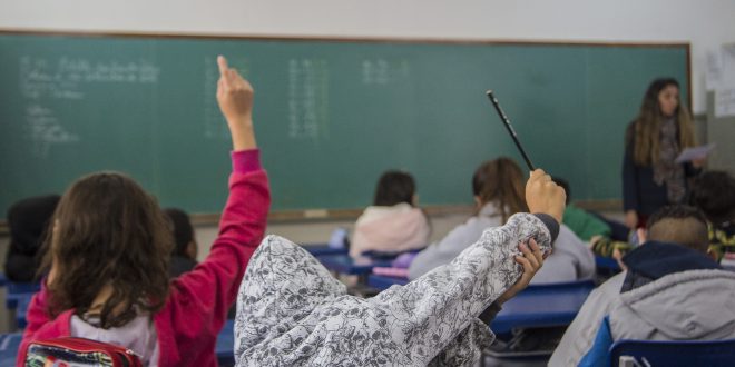 alunos vistos de costas e levantando suas mãos para tirarem dúvidas em sala de aula da Escola Municipal Edith dos Santos Silva