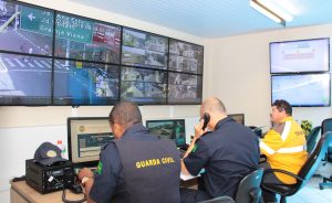 dois guardas civis e um agente de trânsito sentados diante de monitores com imagens de câmeras de segurança e monitoramento de trânsito pela cidade