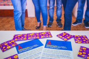 preservativos e folders dispostos sobre mesa com pernas femininas visíveis ao fundo, em segundo plano