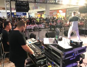 um cantor se apresentando num palco no plaza shopping carapicuíba com um operador de audiovisual ao lado e o público ao fundo