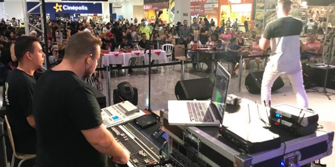 um cantor se apresentando num palco no plaza shopping carapicuíba com um operador de audiovisual ao lado e o público ao fundo