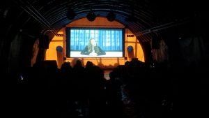 uma tenda arqueada cheia de pessoas vendo um filme projetado numa tela