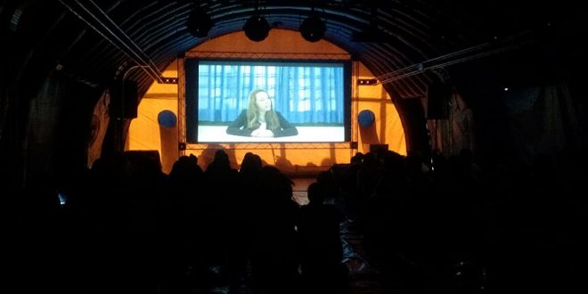 uma tenda arqueada cheia de pessoas vendo um filme projetado numa tela