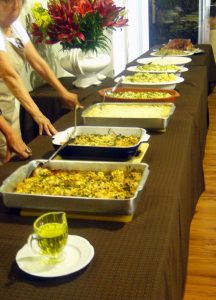 diversas travessas com comidas portuguesas dispostas em uma mesa com mulheres prontas para servir convidados